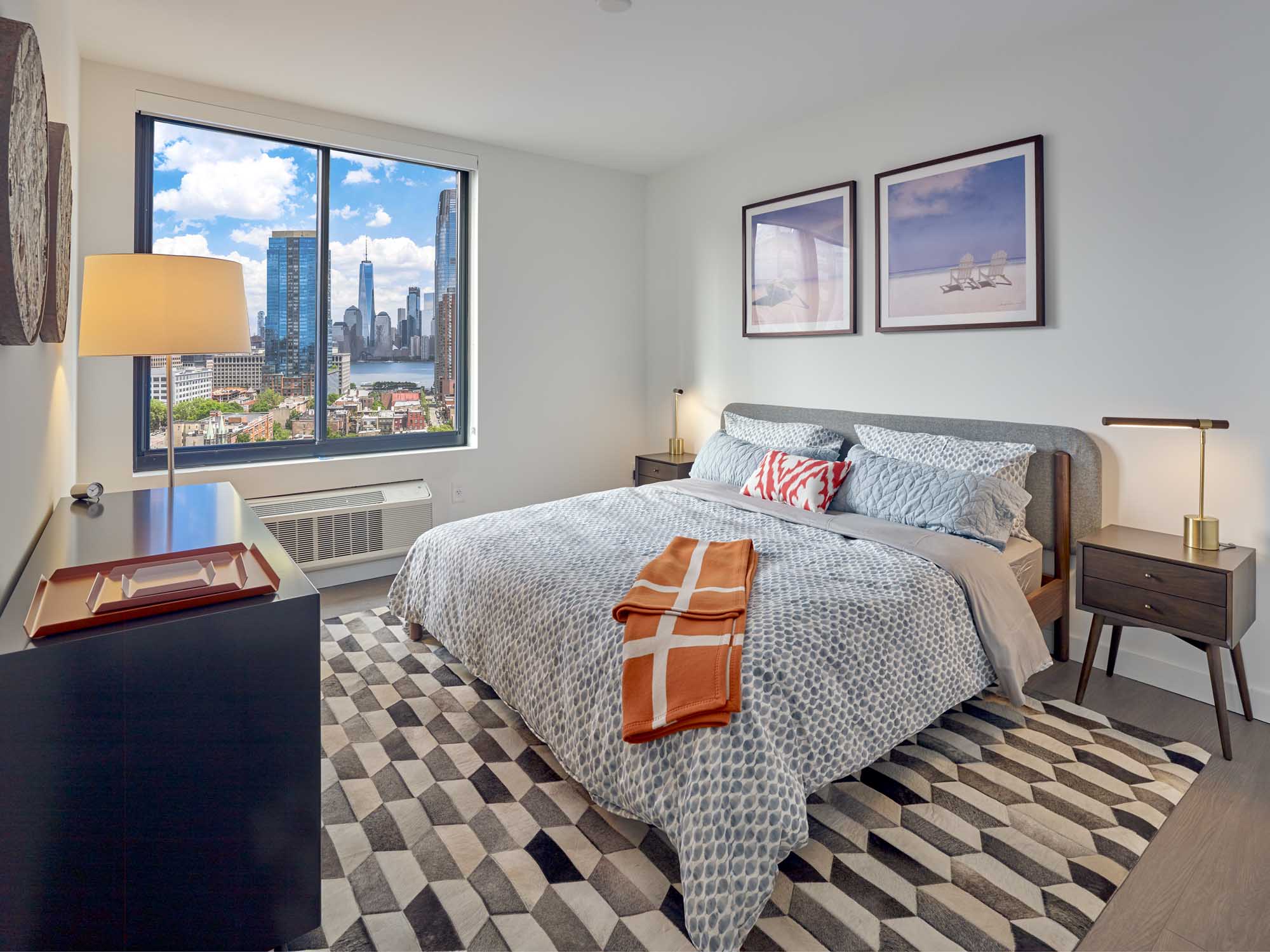 Large bedroom with modern decor, bed, two night tables and large dresser. City views through window.